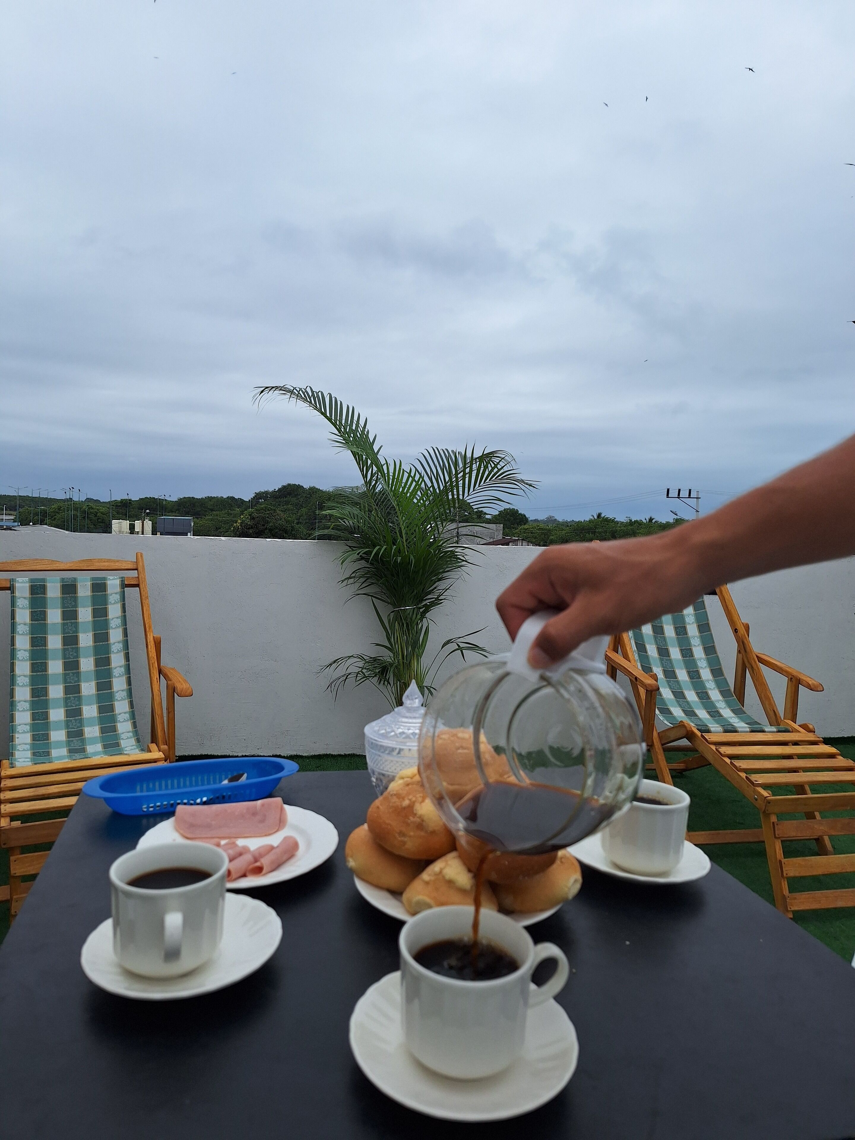 A table with a coffee pot, cups, and pastries, set outdoors with a view of the sky and greenery.