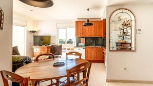 A dining area with a wooden table and chairs, a TV, and a kitchen with wooden cabinets and a mirror. - Sant Genis 19.2 Top floor apartment with terrace (Llança)