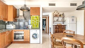 A modern kitchen with wooden cabinets, a built-in oven, a microwave, and a washing machine. There is a dining area with a wooden table and chairs. - Sant Genis 19.2 Top floor apartment with terrace (Llança)