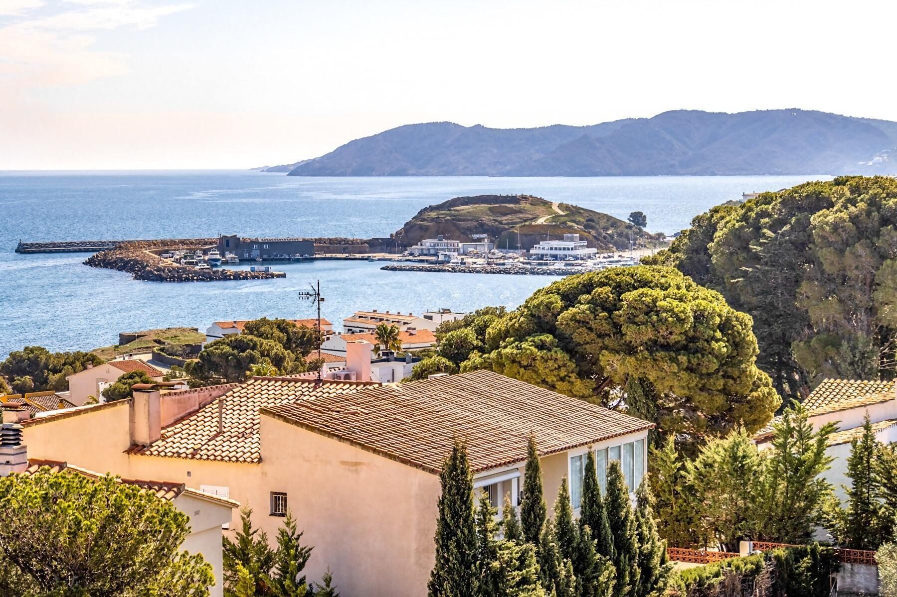 A coastal town with terracotta-roofed buildings, a marina, and a hill with a path leading to a small settlement.