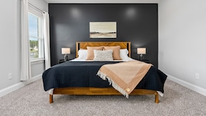 A bedroom with a dark wall, a wooden bed frame, two bedside tables with lamps, a window with curtains, and a painting above the bed.