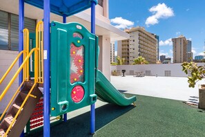 A playground with a slide and climbing structure, set against a building with a clear view of surrounding high-rise buildings.
