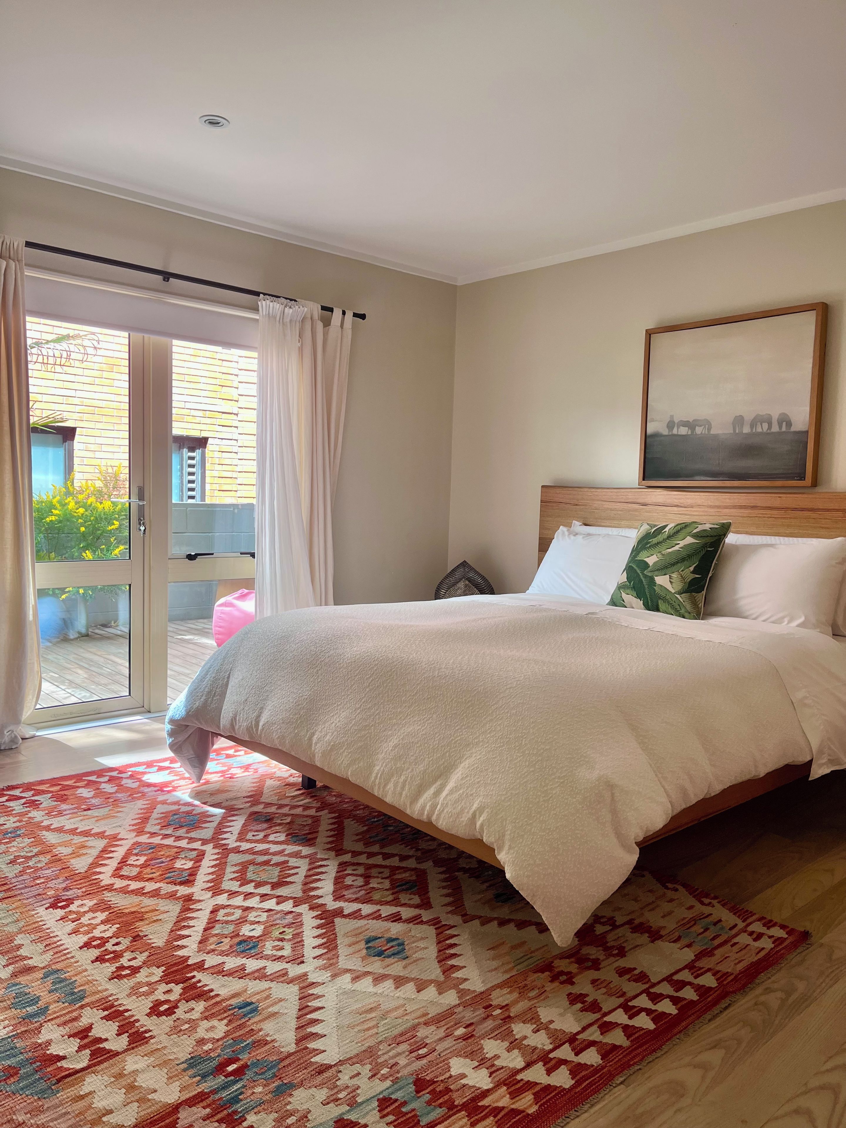 A bedroom with a bed, a patterned rug, a painting, and a view through a glass door.