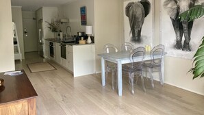 Cocina moderna con armarios blancos, un refrigerador de acero inoxidable y una zona de comedor con mesa de madera y sillas transparentes.