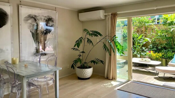 Una sala de estar moderna con una mesa de vidrio, sillas transparentes, una planta grande en maceta y un amplio ventanal corredizo que da a un jardín.