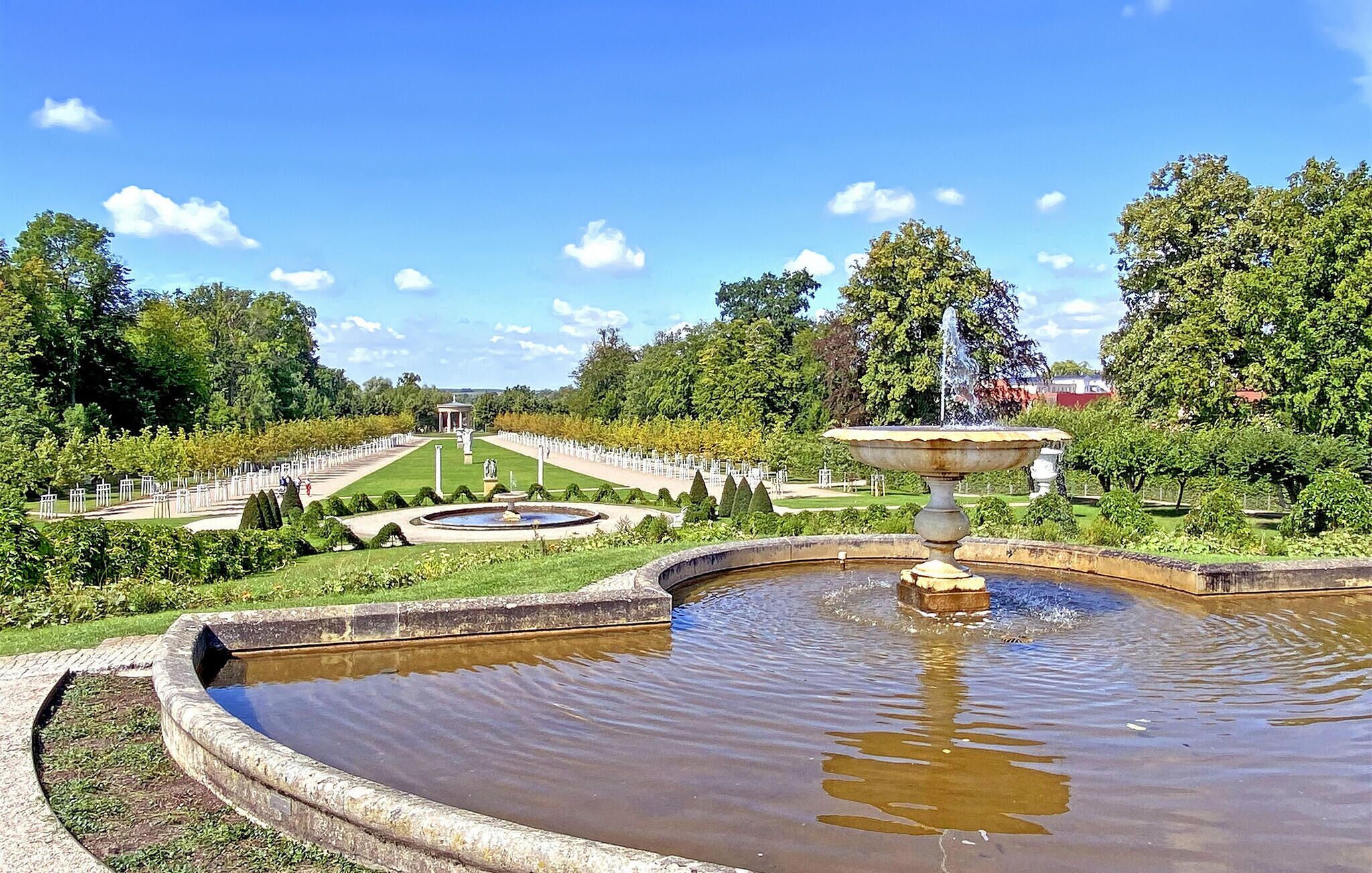 Ein formeller Garten mit einem zentralen Brunnen, gepflegten Rasenflächen und einem klassizistischen Gebäude im Hintergrund.