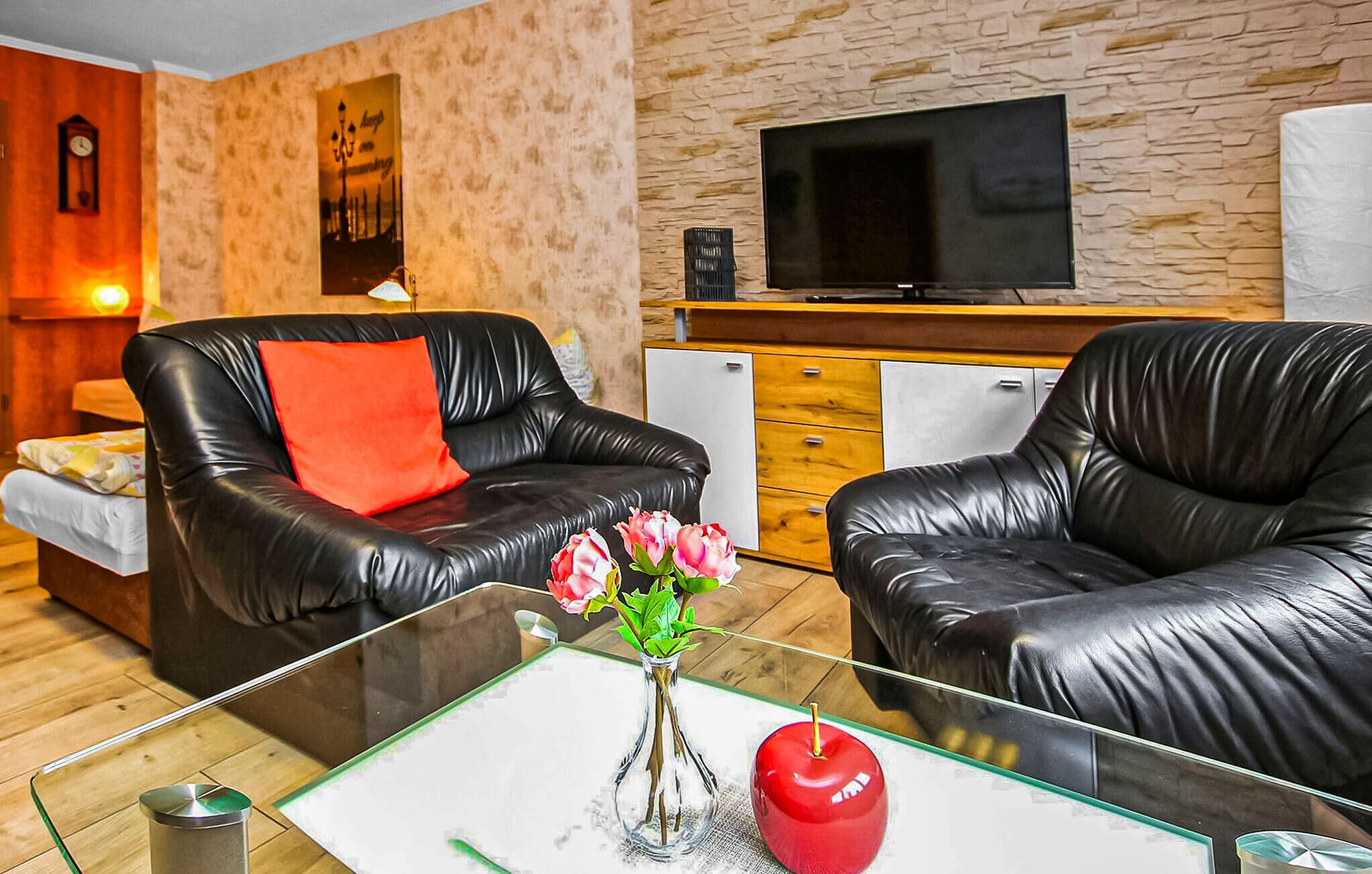 Ein Wohnzimmer mit zwei schwarzen Ledersofas, einem Glastisch mit Blumen und einem roten Apfel, einem Flachbildfernseher an der Wand und einem kleinen Kühlschrank.