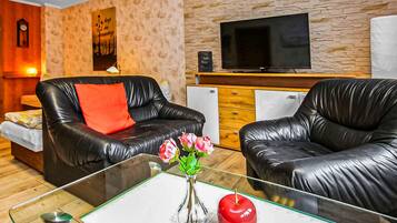 Ein Wohnzimmer mit zwei schwarzen Ledersofas, einem Glastisch mit Blumen und einem roten Apfel, einem Flachbildfernseher an der Wand und einem kleinen Kühlschrank.