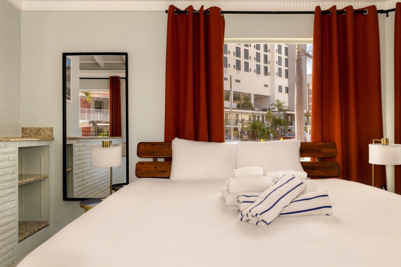 Ein Hotelzimmer mit einem großen Bett, einem Spiegel, einem Fenster mit Vorhängen und Blick auf die Stadt.