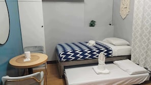 Ein kleiner, ordentlicher Raum mit einem Bett, einem Sessel, einem kleinen Tisch und einer Topfpflanze.