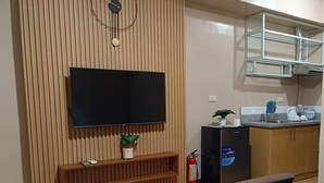 Habitación de hotel con cama, televisión montada en una pared de madera, una pequeña estantería con una planta, extintor y kitchenette con lavaplatos y microondas.
