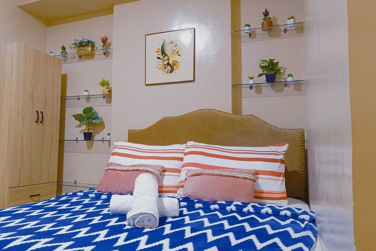 Un dormitorio con cama, almohadas a rayas, una toalla enrollada, un armario de madera y estantes con plantas.