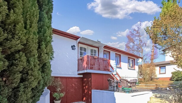 Una casa de dos pisos con fachada roja y blanca, un balcón con mobiliario de mimbre y un pequeño jardín.