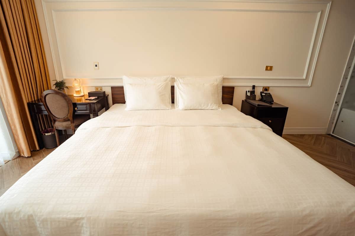 Cama bem arrumada com cabeceira, mesa de cabeceira com abajur e um espelho.