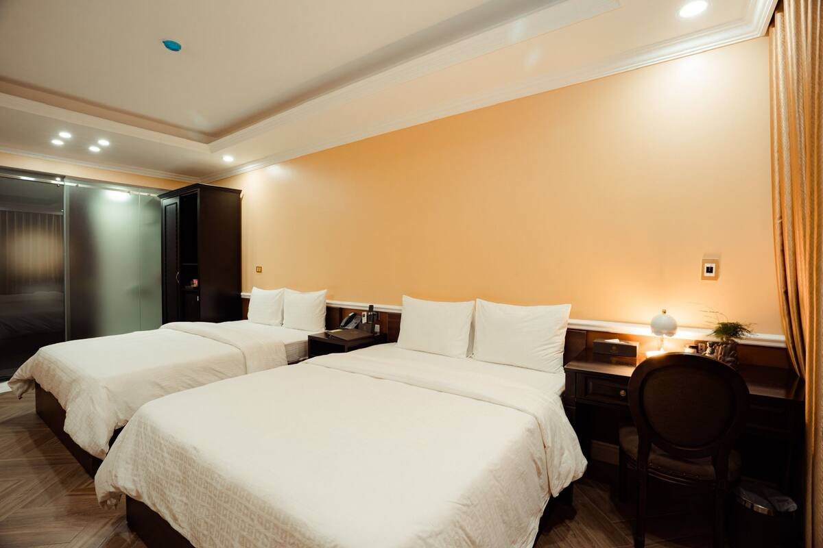 Quarto de hotel com duas camas, uma escrivaninha, uma cadeira e uma mesa de cabeceira com abajur.
