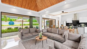 A modern living room with a wooden ceiling, grey sofas, a coffee table, and a view of a garden and kitchen area. - Luxury Balinese Private Pool Villa-KBR12 (Koh Samui)