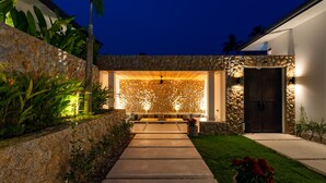 A modern house with a stone wall, a wooden deck, and a well-lit entrance. - Luxury Balinese Private Pool Villa-KBR12 (Koh Samui)