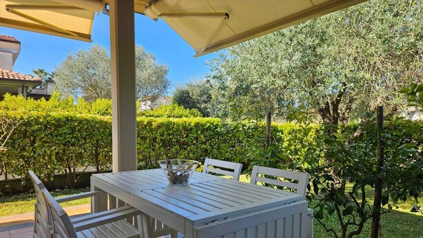 An outdoor patio with a table and chairs, surrounded by greenery and a clear sky.