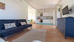 A modern living room with a dark sofa, a white armchair, and a beige rug. A kitchen with white cabinets and a blue cabinet is visible in the background.
