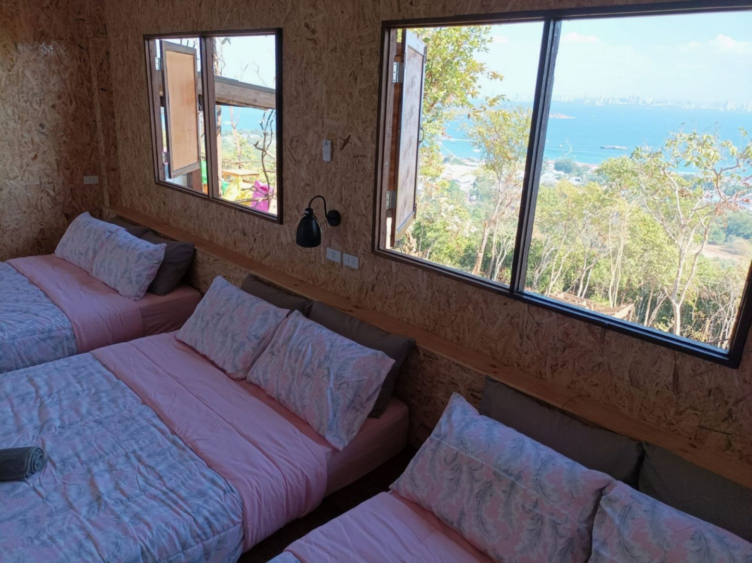 A room with three beds, large windows, and a view of the sea and trees.