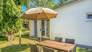 A patio with a wooden table and wicker chairs, an umbrella, and a white building with a window.