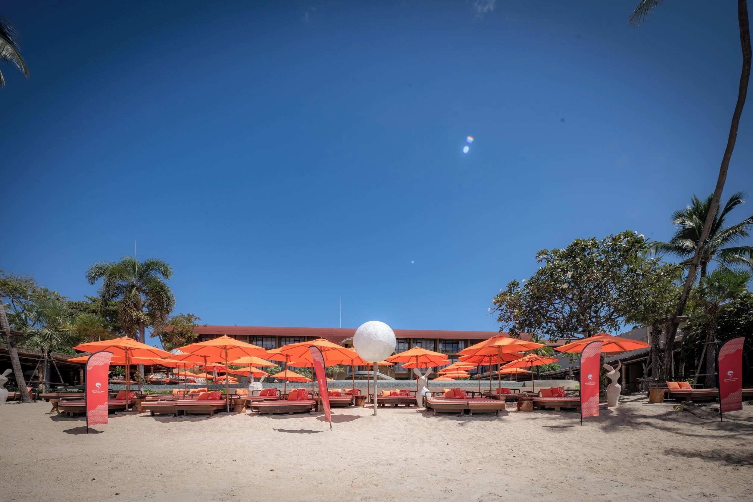 A beach with rows of orange umbrellas and lounge chairs, palm trees, and a building in the background.