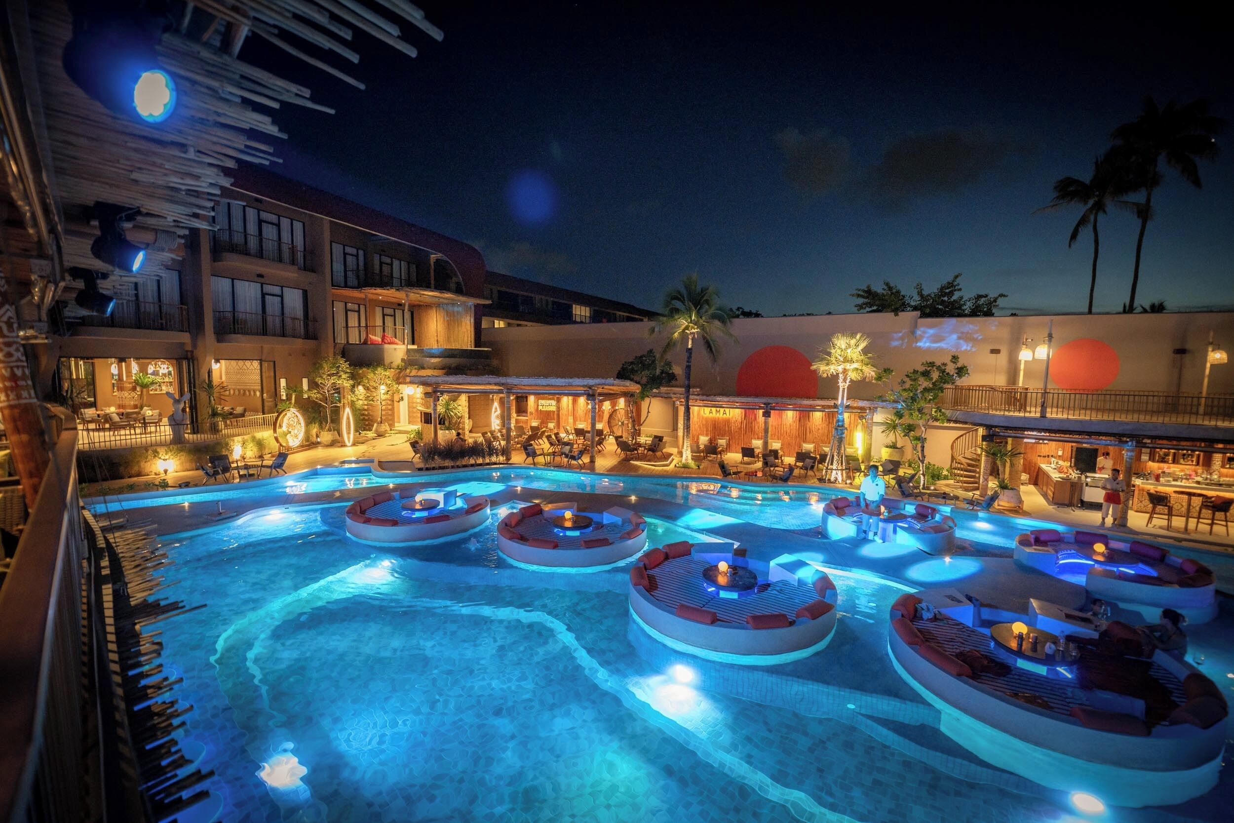 A pool area with multiple circular seating areas around the pool, a bar area with outdoor seating, and a building with multiple floors and balconies.
