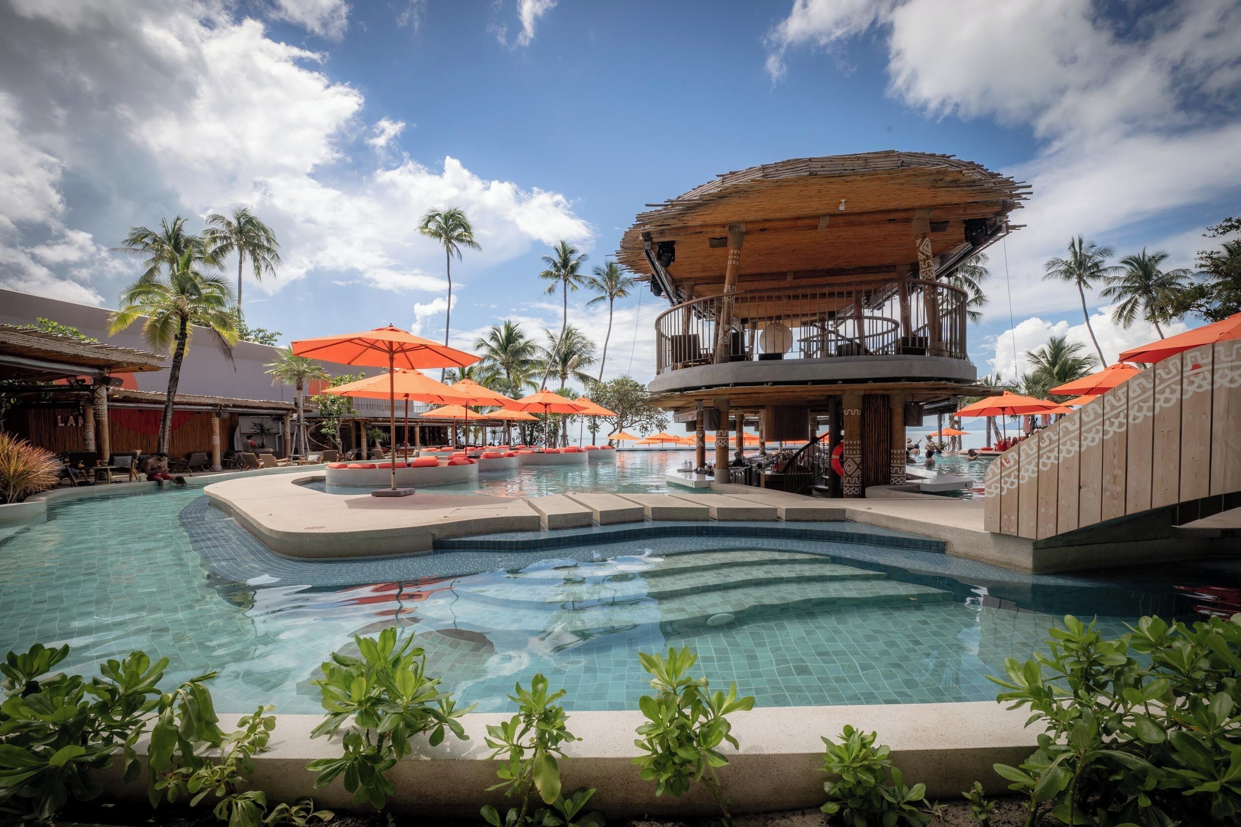 A pool area with a wooden structure, orange umbrellas, and palm trees.
