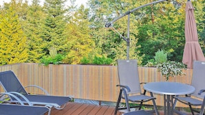 Eine Außenpatio mit Holzdeck, Metallstühlen, einem runden Tisch und einer Pergola mit Sonnenschirm.