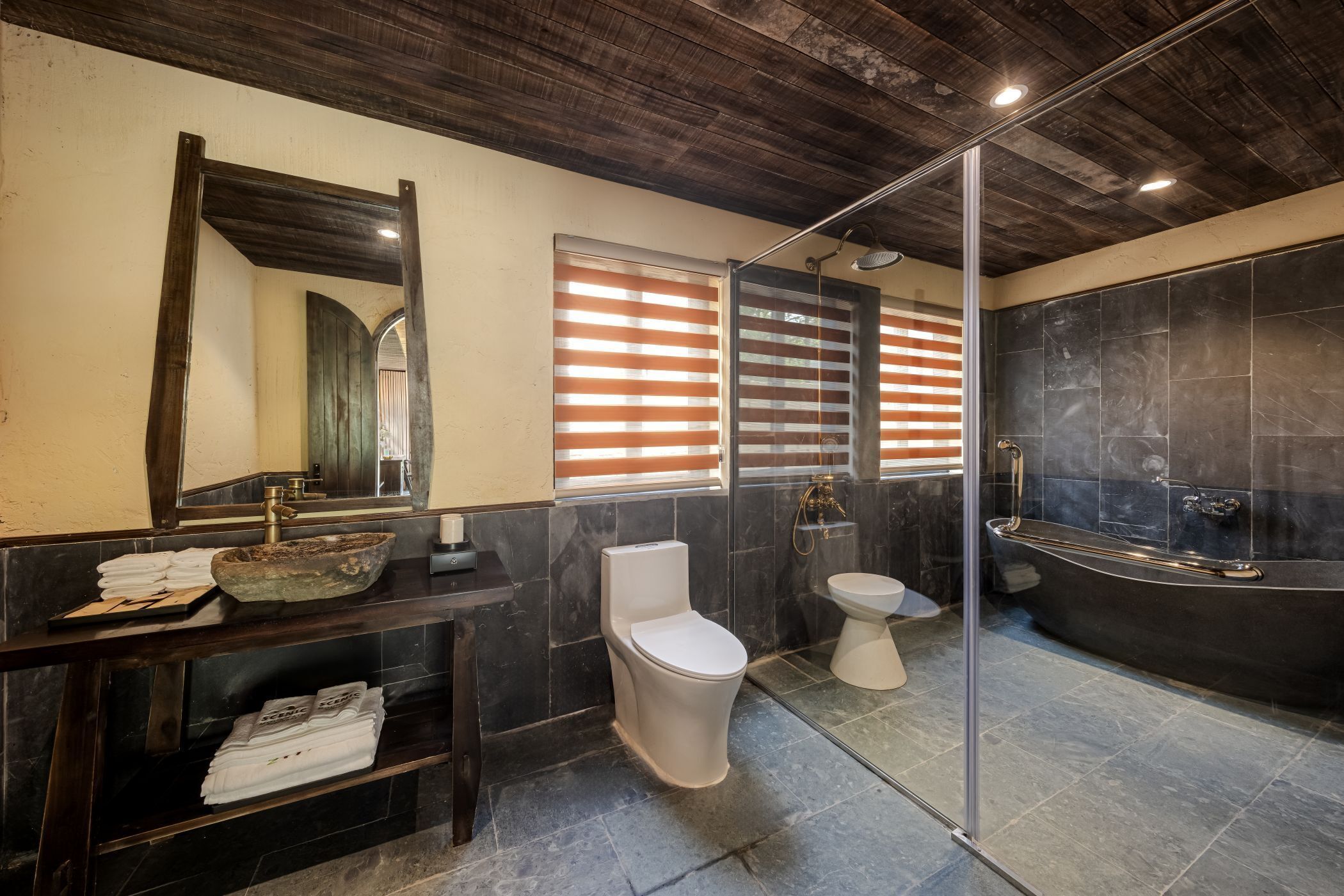 A modern bathroom with a dark wood ceiling, a large mirror, a sink with a bowl, a toilet, and a bathtub.