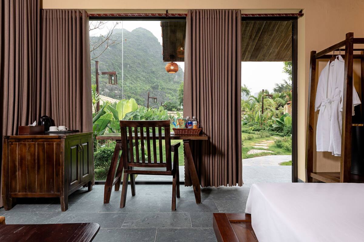 A room with a bed, a wooden desk, a chair, and a view of a lush garden and mountains.