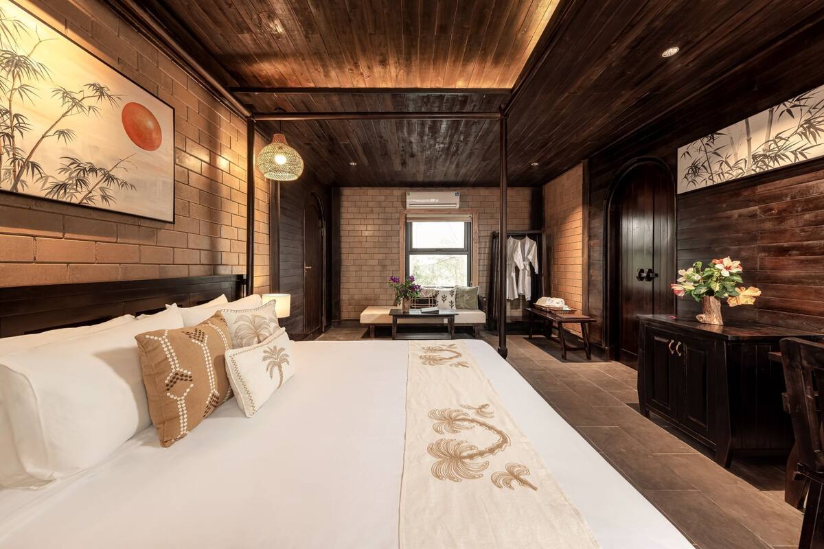 A hotel room with a large bed, wooden ceiling, and a brick wall. There is a window, a bench, and a small table with a vase of flowers.