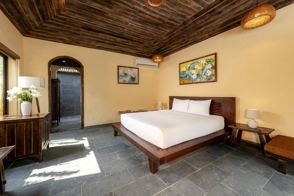 A bedroom with a wooden bed, a desk with a lamp, a bench, and a painting on the wall.