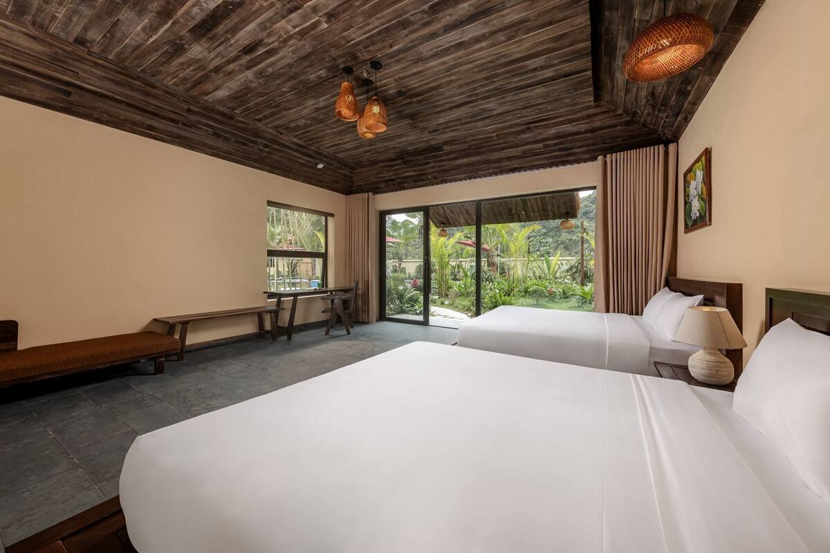 A hotel room with two beds, a wooden ceiling, large windows, a bench, and a table.