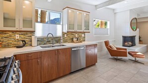 A modern kitchen with wooden cabinets, a dishwasher, and a large island. There is a fireplace and a seating area with a cushioned chair and ottoman.