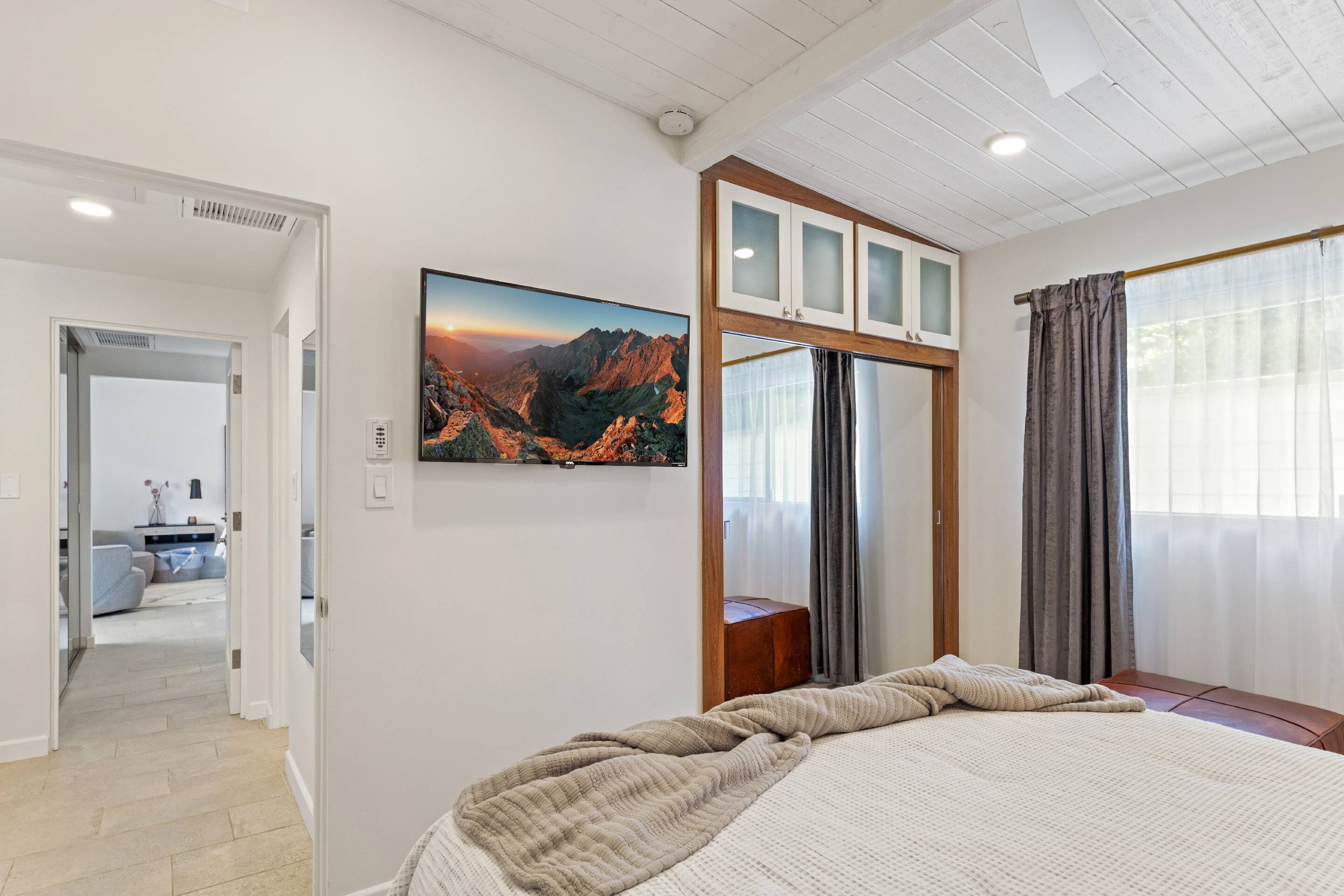 Un dormitorio con cama, un ventanal con cortinas, un cuadro colgado de una montaña y una puerta que da a otra habitación.