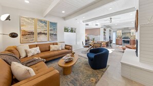 A spacious living room with a large brown leather sofa, a blue armchair, and a wooden coffee table. There are framed artworks on the wall and a kitchen area in the background.