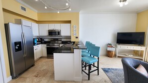 A compact kitchen with a refrigerator, microwave, and oven. There is a small dining area with a table and four chairs. A television is mounted on a stand to the right. - Direct Beachfront Condo Near Pier Park! 2BD+Bunk 3BA, 2 Free Parking! (Panama City Beach)