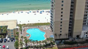A hotel with a swimming pool surrounded by palm trees, a beach with umbrellas, and a parking lot. - Direct Beachfront Condo Near Pier Park! 2BD+Bunk 3BA, 2 Free Parking! (Panama City Beach)