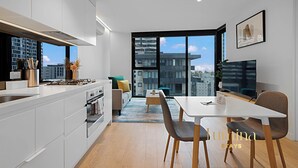 Une cuisine moderne dotée d’un îlot central, d’un coin repas avec une table et des chaises blanches, et d’une grande fenêtre offrant une vue sur la ville.