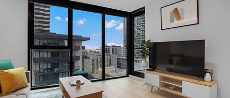 A modern living room with a large TV, a sofa, a coffee table, and a view of the city through large windows.