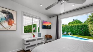 Ein Schlafzimmer mit einem großen Bett, einem Fernseher, einem Schminktisch mit Spiegel und einem Fenster mit Blick auf Pool und Garten.