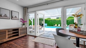 Ein modernes Wohnzimmer mit Holzboden, einem Essbereich mit Tisch und Stühlen, einer großen Schiebeglastür, die zu einem Pool führt, und gerahmten Kunstwerken an der Wand.
