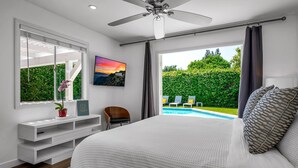 Ein Schlafzimmer mit einem großen Bett, einem Deckenventilator, einem Schreibtisch mit Stuhl, einem Fenster mit Blick auf Pool und Grünflächen sowie einem Bild an der Wand.