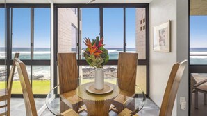 Un coin repas avec une table en verre, des chaises en bois, un ventilateur de plafond et une vue sur l’océan grâce à de grandes fenêtres.