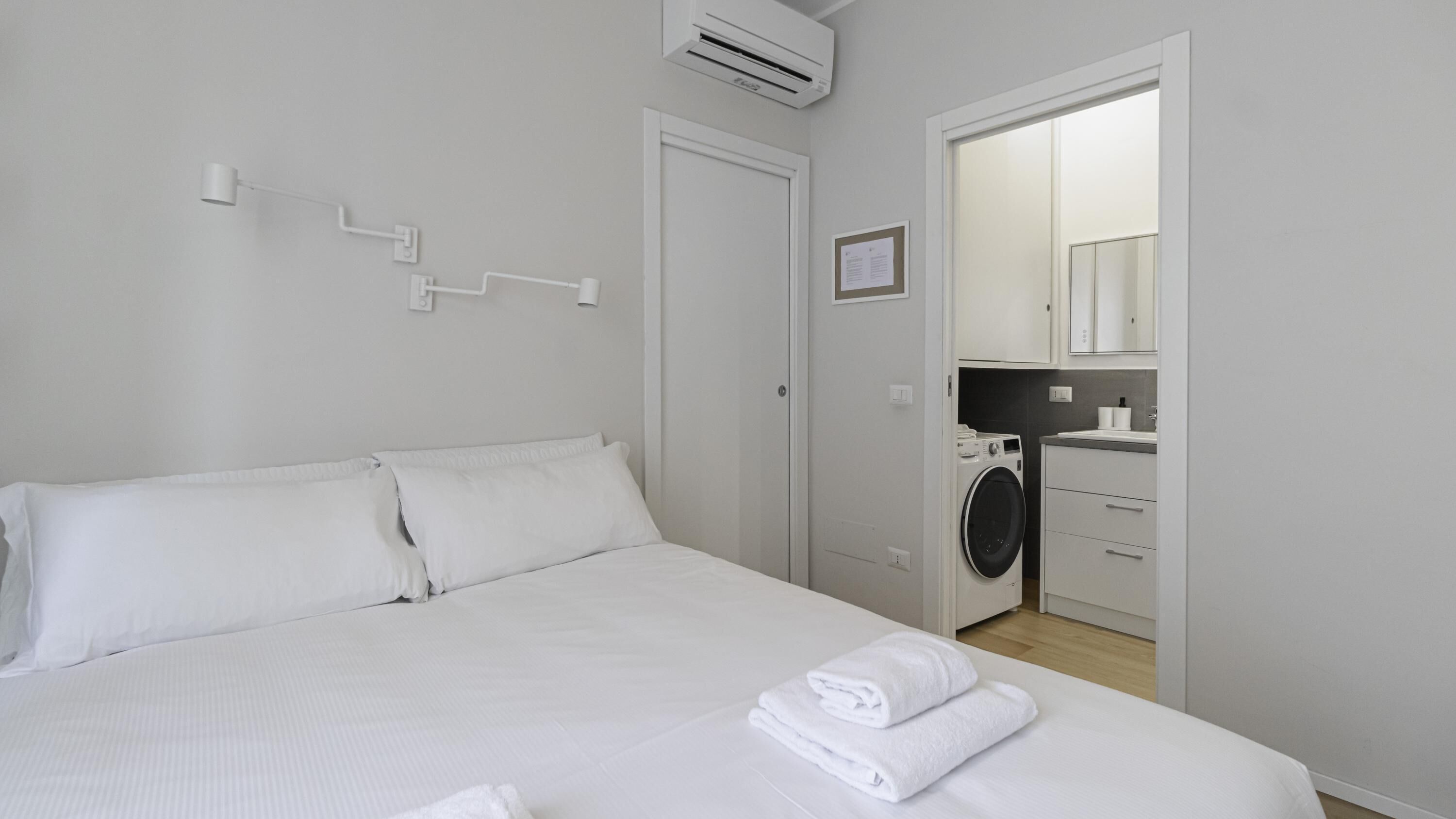 Une chambre à coucher avec un lit, du linge de lit blanc, un lave-linge et une salle de bain visible à travers une porte ouverte.