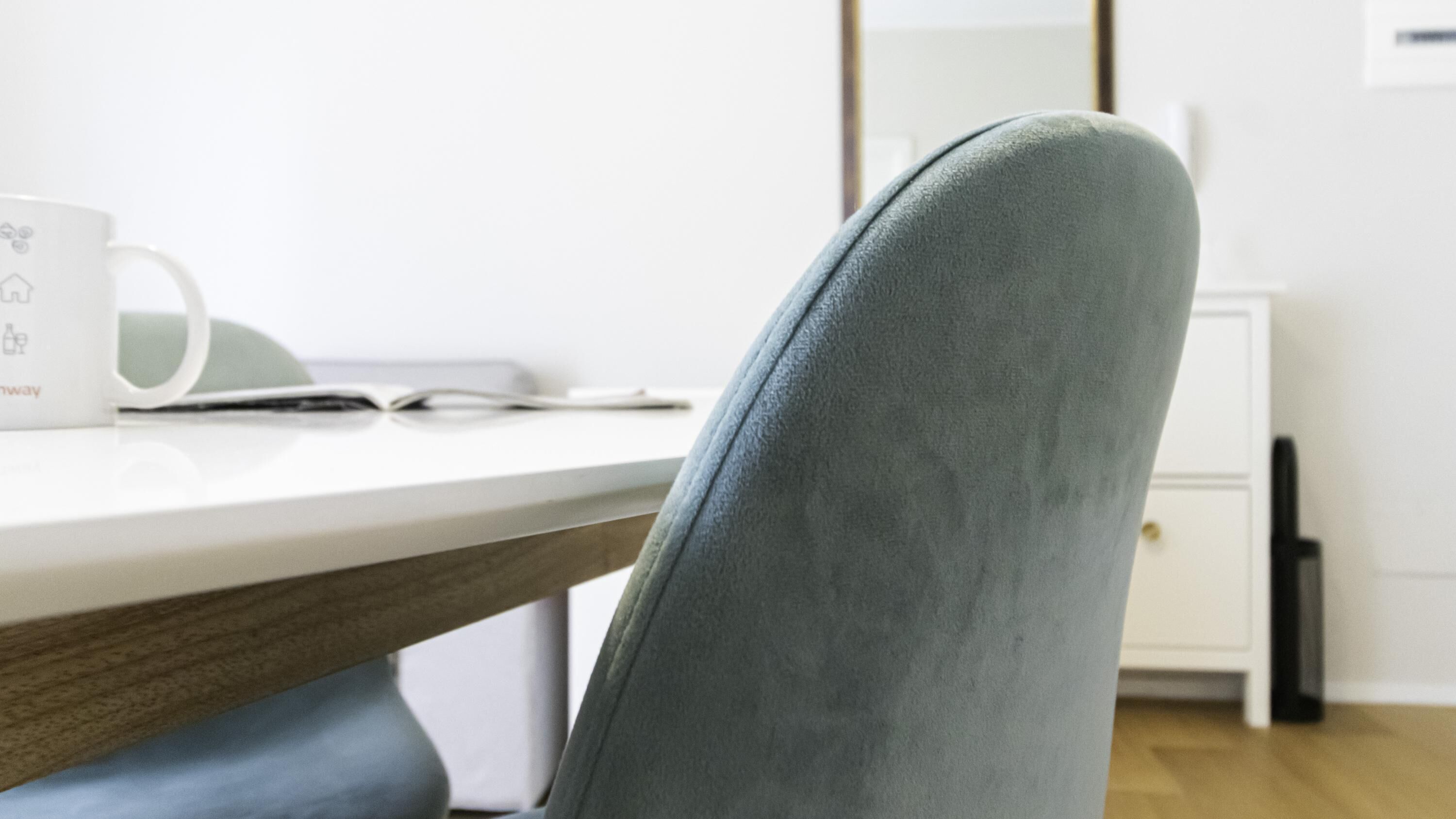 Une chaise de bureau moderne, recouverte d’un tissu gris, un bureau blanc avec une tasse, et un meuble à tiroirs blanc en arrière-plan.