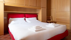 Un lit bien fait, avec du linge de lit blanc et une tête de lit rouge, une chaise en bois et une armoire en bois.