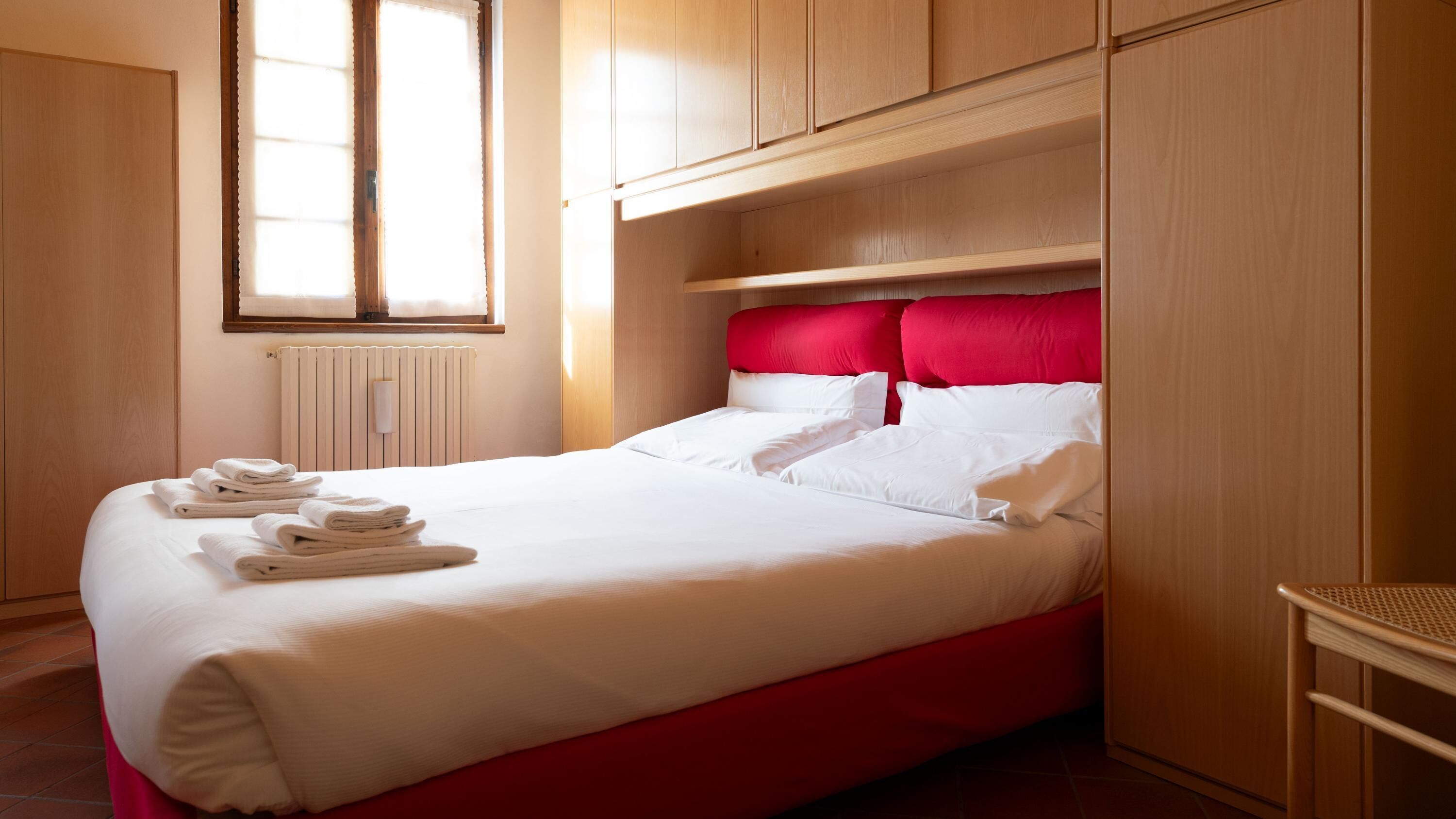 A neatly made bed with white linens and red headboard, a window with blinds, and wooden cabinetry.