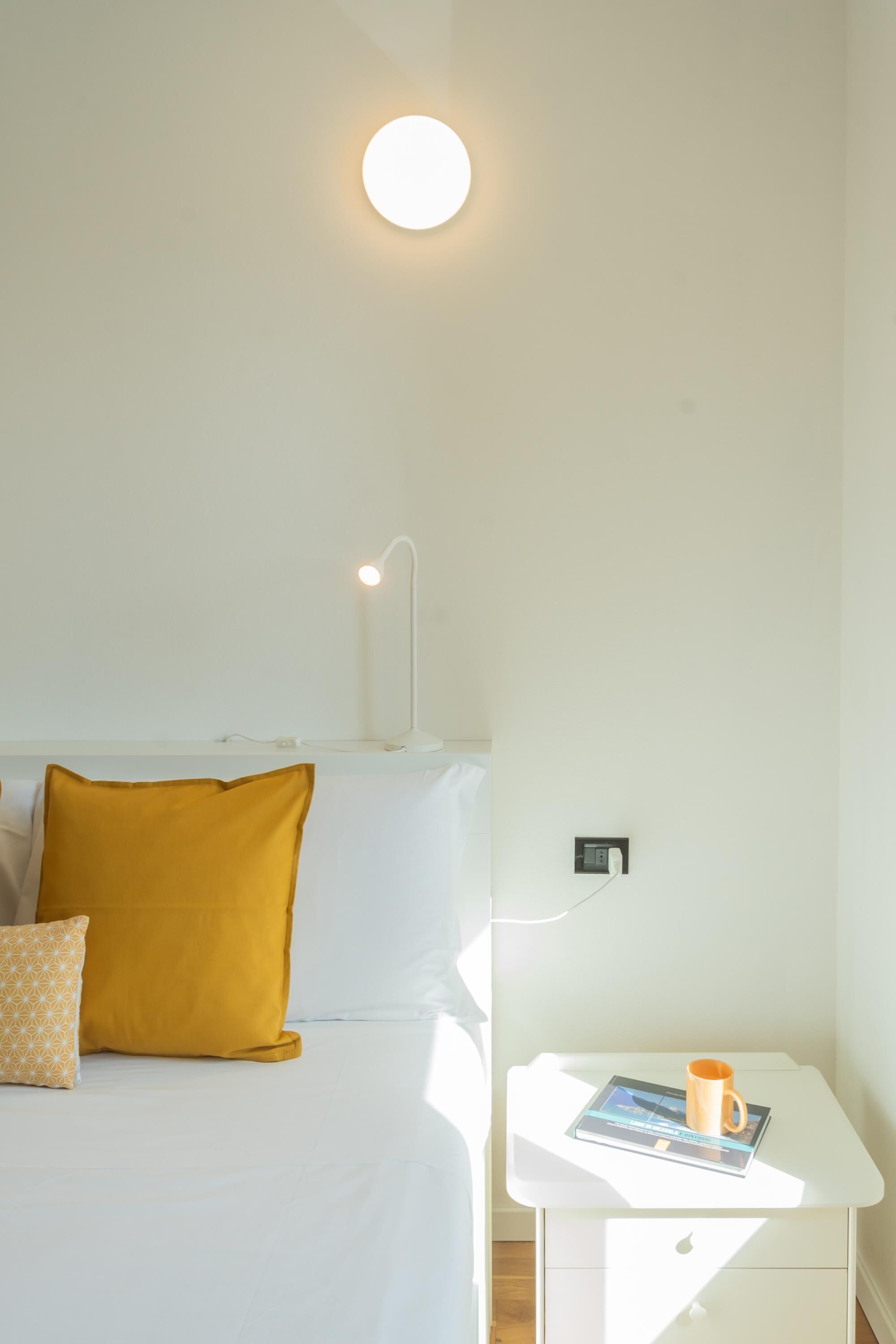A neatly arranged hotel room with a bed, a bedside table with a book and a mug, and a wall-mounted light fixture.